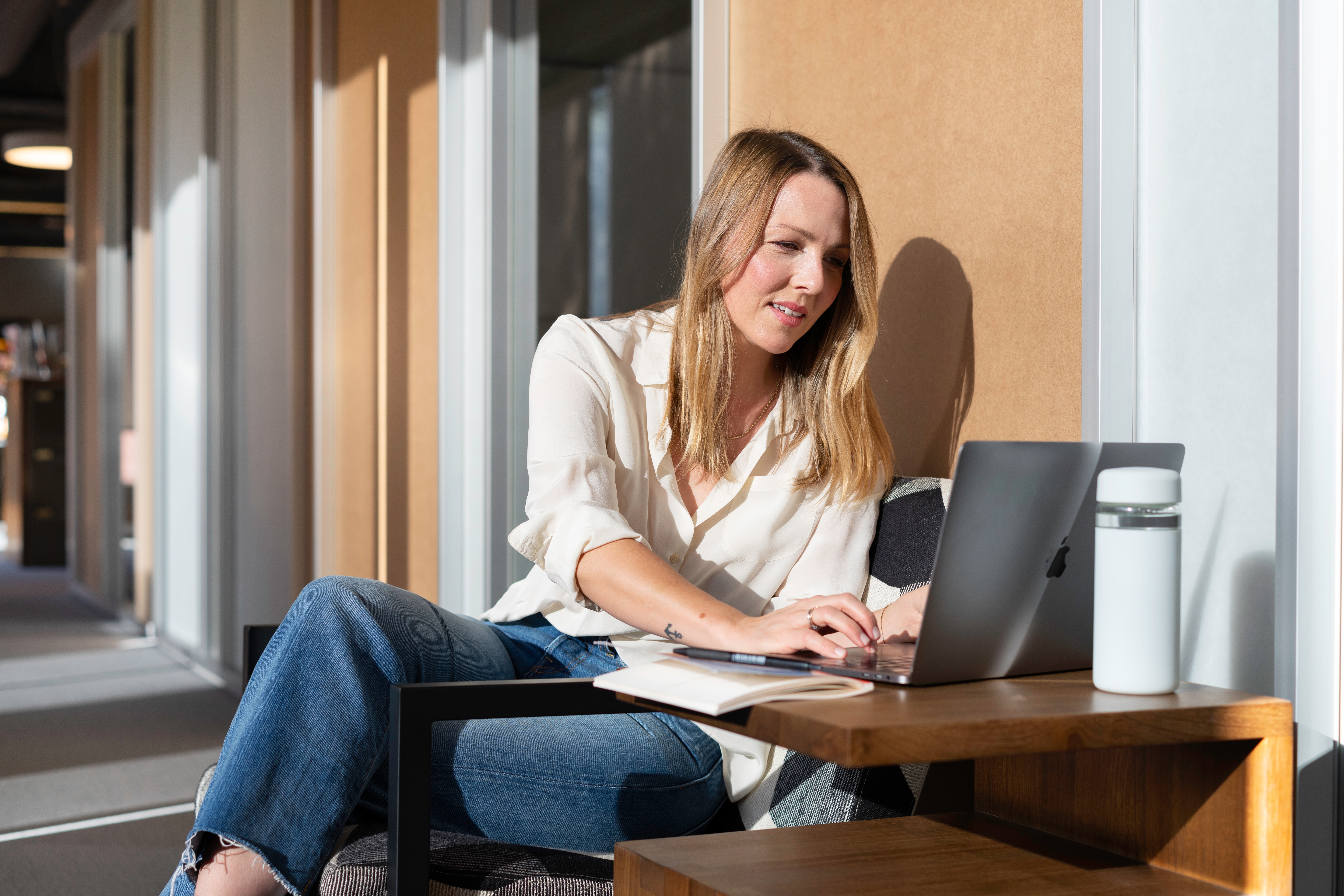 A blonde woman sitting at a low table using a laptop. She is wearing a white shirt and jeans